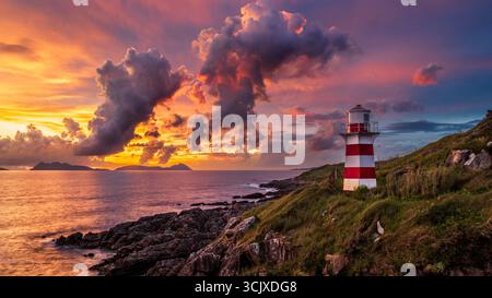 Ein rot-weiß gestreifter Leuchtturm steht auf einer felsigen, grünen Klippe mit Blick auf das ruhige Meer. Der Himmel darüber ist ein atemberaubendes Spektakel eines Sonnenuntergangs, mit Stockfoto