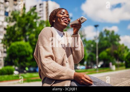 Die junge glückliche schwarze Frau benutzt ein Smartphone. Sie lacht, während sie Sprachnachrichten aufnimmt. Stockfoto