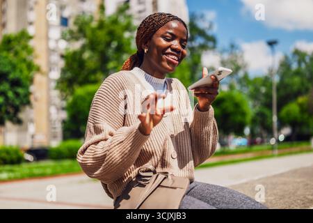 Die junge glückliche schwarze Frau benutzt ein Smartphone. Sie lacht, während sie Sprachnachrichten aufnimmt. Stockfoto