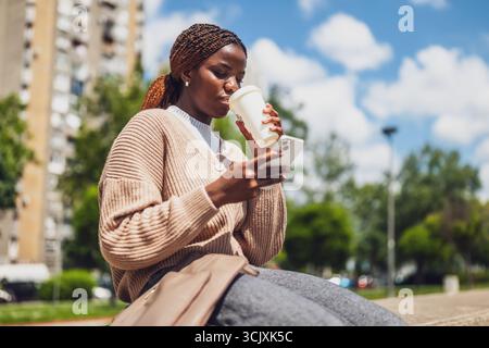 Nachdenkliche junge schwarze Frau, die in einem Park sitzt und auf ihrem Smartphone SMS schreibt, während sie an einem sonnigen Tag einen Kaffee genießt. Stockfoto