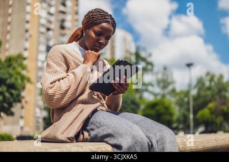 Afroamerikanerin in ihren 20ern sieht besorgt und unzufrieden aus, als sie mit ihrem Tablet interagiert. Sie sitzt im Freien und spiegelt den Druck des ci wider Stockfoto