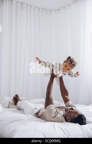 Glücklicher afrikanischer Vater hebt seine niedliche, birassische Tochter auf, während er im hellen weißen Zimmer auf dem Bett liegt. Das Konzept der interrassischen Familie. Stockfoto