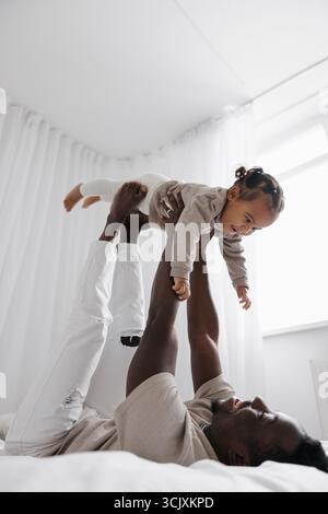 Glücklicher afrikanischer Vater hebt seine niedliche, birassische Tochter auf, während er im hellen weißen Zimmer auf dem Bett liegt. Das Konzept der interrassischen Familie. Stockfoto