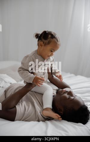 Glücklicher afrikanischer Vater, der auf dem Bett liegt und mit seiner niedlichen, birassischen Tochter in einem hellen weißen Zimmer spielt. Das Konzept der interrassischen Familie. Stockfoto