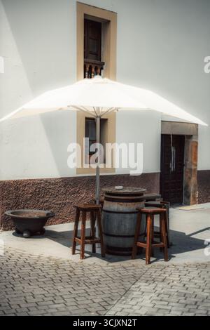 Rustikale Sitzmöbel im Freien mit Holzfasstisch, Vintage-Hockern und einem großen weißen Regenschirm vor einer traditionellen Stuckwand, die einen Saibling schafft Stockfoto