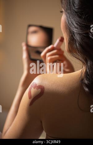 Die weibliche Figur trägt Lippenstift auf, während sie in einen Spiegel blickt. Sie zeigt ein Herzdesign auf ihrer Schulter, das Schönheit und persönliche Pflege in Ruhe betont Stockfoto