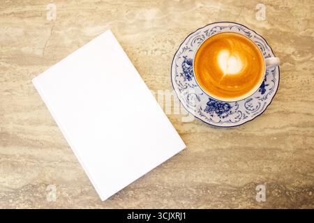 Kaffeetasse mit herzförmiger Latte Art, die neben einem leeren Buch auf einem Marmortisch platziert wird, schafft eine warme und einladende Szene zum Entspannen und Inspi Stockfoto