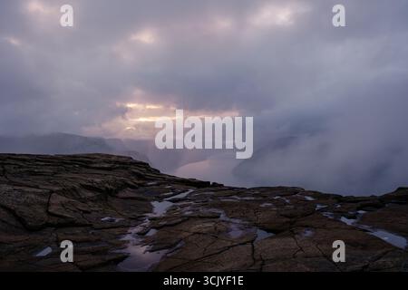 Erleben Sie die atemberaubende Schönheit der Preikestolen Cliff in Norwegen, während die Sonne hinter der zerklüfteten Landschaft untergeht und einen mystischen Glanz über den darunter liegenden Fjord und den umliegenden Nebel wirft. Stockfoto