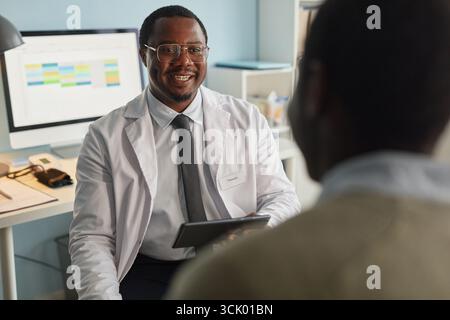 Schwarzer Mann im mittleren Alter, Arzt lächelt, während er den Patienten in der Praxis berät Stockfoto