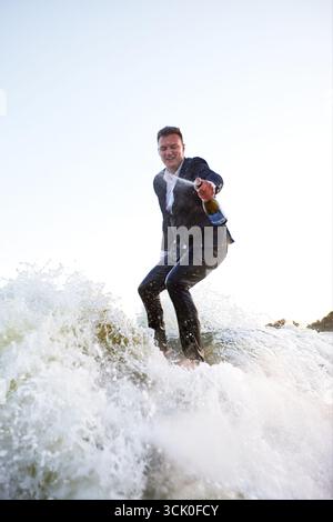 Junger Mann im klassischen Anzug mit Champagnerflasche fährt auf einem Wakeboard auf dem See in der Nähe der Stadt. Happy Clerk ist aus einem stickigen Büro geflohen, um aktive sp Stockfoto