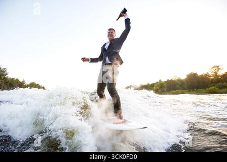 Junger Mann im klassischen Anzug mit Champagnerflasche fährt auf einem Wakeboard auf dem See in der Nähe der Stadt. Happy Clerk ist aus einem stickigen Büro geflohen, um aktive sp Stockfoto
