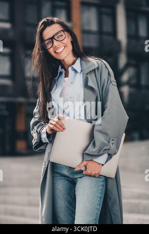 Junge Frau in modernem Trenchcoat posiert draußen mit Laptop vor städtischem Hintergrund, während sie tagsüber vertikales Porträt hält Stockfoto