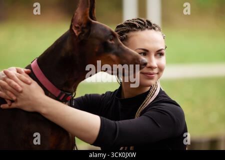 Eine junge Frau mit stylischen geflochtenen Haaren und einem schwarzen Oberteil umarmt einen Doberman mit einem rosa Kragen. Die lebhafte Umgebung im Freien bietet einen grünen Hintergrund Stockfoto