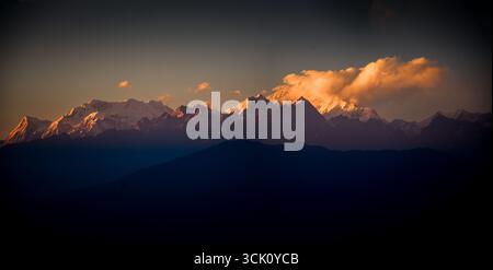 Majestätische Berggipfel, die mit Schnee bedeckt sind, erstrahlen in den goldenen Tönen des Sonnenuntergangs und bilden einen wunderschönen Kontrast zur dunklen Landschaft im Vordergrund Stockfoto