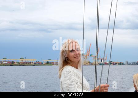Eine Frau im mittleren Alter mit blonden Haaren, in einem weichen Pullover gekleidet, steht auf einem Boot, umgeben von ruhigem Wasser und einem malerischen Himmel, und fängt eine Momen ein Stockfoto