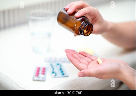 Frau gießt Pillen aus der bernsteinfarbenen Flasche in ihre Hand mit Blisterpackungen und einem Glas Wasser in der Nähe, Konzept-Hausmedikation und tägliche Gesundheit Routine Stockfoto