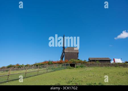 Windmühle mit Zaun und Himmel. Eine hölzerne Windmühle steht auf einem Hügel mit einem Zaun darunter. Helle Blumen und ein tiefblauer Himmel runden die ländliche Szene ab. Stockfoto