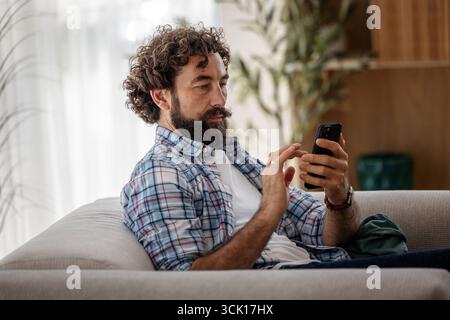 Gutaussehender Mann, der sich auf dem Sofa in seiner Wohnung entspannt und mit dem Telefon eine SMS an die Frau geschickt hat, mit fokussiertem Gesichtsausdruck Stockfoto