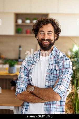 Ein Mann mit lockigen Haaren und Bart steht in einer hellen Küche und lächelt selbstbewusst, während er ein kariertes Hemd über einem weißen T-Shirt trägt Stockfoto