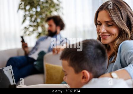 Mutter genießt entspannte Momente mit ihrem Sohn auf dem Sofa, während der Vater im Hintergrund ist und mit seinem Handy in ihrem gemütlichen Wohnzimmer beschäftigt ist. Stockfoto