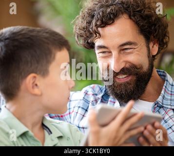 Vater und Sohn teilen freudige Momente, während sie mit dem Tablet in entspannter, natürlicher Umgebung interagieren. Sie lächeln einander an Stockfoto
