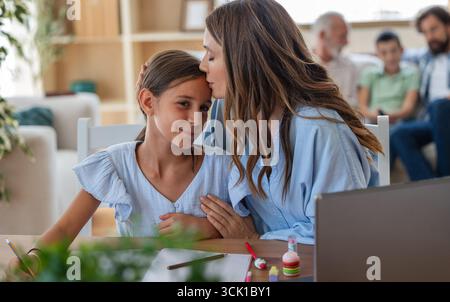 Mutter küsst ihre Tochter auf die Stirn, während sie zu Hause kreative Aktivitäten und Hausaufgaben macht. Familienmitglieder sind im Hintergrund sichtbar Stockfoto