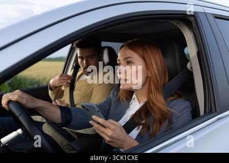 Eine junge Geschäftsfrau, die versucht, beim Autofahren vorsichtig ein Telefon zu benutzen, neben ihr schaut ein Mann ängstlich zu. Stockfoto