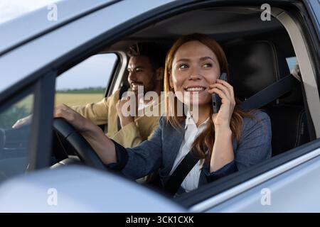Fröhliche Frau, die auf einem Mobilgerät spricht, während sie ein Lenkrad hält und aus einem Fenster blickt, lächelt der Mann neben ihr im Auto. Mult Stockfoto