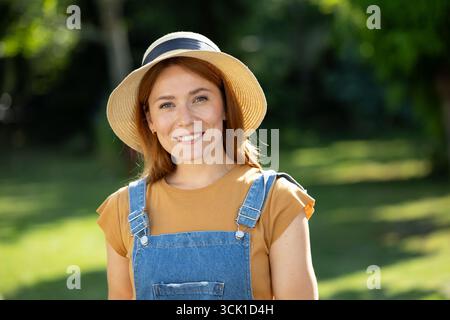 Porträt einer jungen Frau steht im sonnigen Garten, lächelt warm, während sie Strohhut und Jeansanzüge trägt, umgeben von Grün Stockfoto
