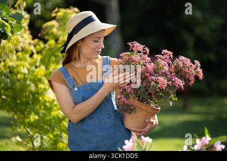 Eine junge Frau, die rosa Blumen in einem Topf beobachtet und sanft lächelt, den sie in ihren Händen hält Stockfoto