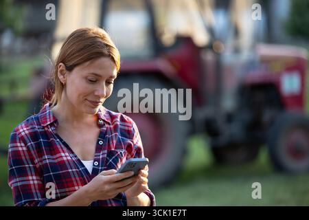 Eine junge Frau auf dem Bauernhof lächelt an ihrem Telefon, während sie vor dem Traktor steht Stockfoto