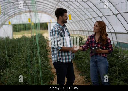 Männliche Bauern und Landwirtschaftsexperten lächeln, während sie sich die Hände schütteln, während sie im Gewächshaus einen Deal machen Stockfoto