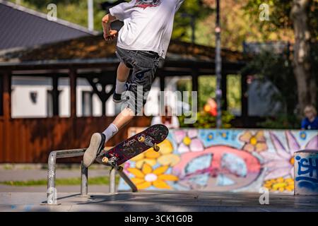 Skater in einem Skatepark, der einen Flip-Trick über eine Box macht, eine Rampe und eine Schiene für Grinding und einen Peace Logo Graffitti auf einer Rampe im Hintergrund Stockfoto