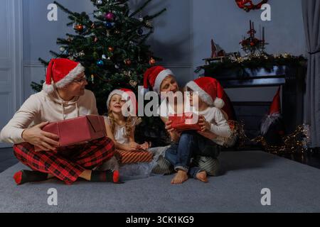 Familie in Ferienhüten sitzt neben dem Baum, hält gewickelte Geschenke Stockfoto