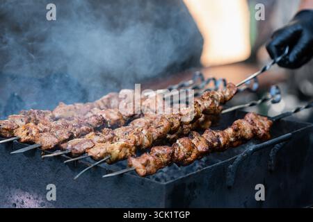 Eine Nahaufnahme einer Person, die Schweinefleischspieße auf einem Grill grillt, mit schwarzem Handschuh Stockfoto