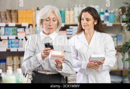 Ältere Frau in der Apotheke scannt QR-Code auf Omega-3-Ergänzung, Frau Apothekerin im Hintergrund Stockfoto