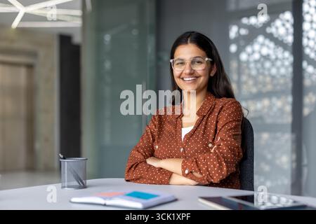 Die junge indische Geschäftsfrau lächelt selbstbewusst mit gekreuzten Armen, während sie an einem modernen Schreibtisch sitzt, und strahlt Professionalität und Ehrgeiz in einer stilvollen Unternehmensumgebung aus Stockfoto
