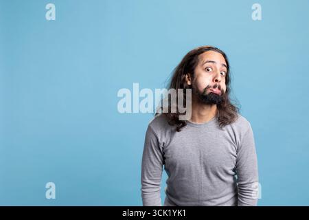 Fröhlicher bärtiger Mann mit langen lockigen Haaren schlägt eine alberne Pose und zeigt eine sorglose Haltung. Spielerisches Verhalten und komisches Gesicht unterstreichen seine humorvolle Persönlichkeit in diesem Studio vor blauem Hintergrund. Stockfoto