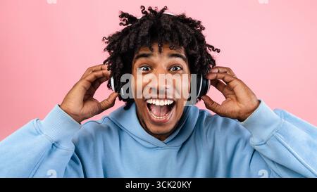 Ein überfröhlicher schwarzer Teenager genießt beliebte Musik im Headset und öffnet den Mund vor Spannung auf rosa Hintergrund Stockfoto