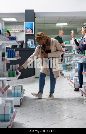 afroamerikanischer Patient überprüft pharmazeutische Artikel in organisierter Drogerie und konzentriert sich dabei auf genaue Dosisinformationen zu Nahrungsergänzungsmitteln zur Selbstversorgung. Arztbesuch. Stockfoto
