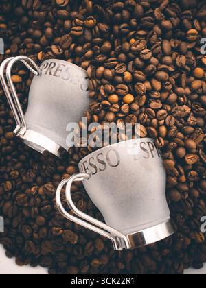 Eine Hochwinkelaufnahme von zwei Espresso-Tassen, die auf einem Bett mit gerösteten Kaffeebohnen liegen. Das Bild hebt die Textur und die tiefbraune Farbe der Bohnen hervor, weiß Stockfoto