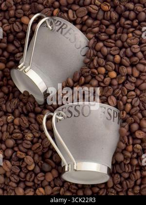 Eine Hochwinkelaufnahme von zwei Espresso-Tassen, die auf einem Bett mit gerösteten Kaffeebohnen liegen. Das Bild hebt die Textur und die tiefbraune Farbe der Bohnen hervor, weiß Stockfoto