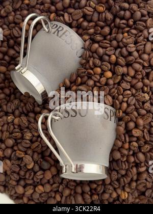 Eine Hochwinkelaufnahme von zwei Espresso-Tassen, die auf einem Bett mit gerösteten Kaffeebohnen liegen. Das Bild hebt die Textur und die tiefbraune Farbe der Bohnen hervor, weiß Stockfoto