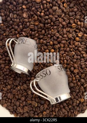 Eine Hochwinkelaufnahme von zwei Espresso-Tassen, die auf einem Bett mit gerösteten Kaffeebohnen liegen. Das Bild hebt die Textur und die tiefbraune Farbe der Bohnen hervor, weiß Stockfoto