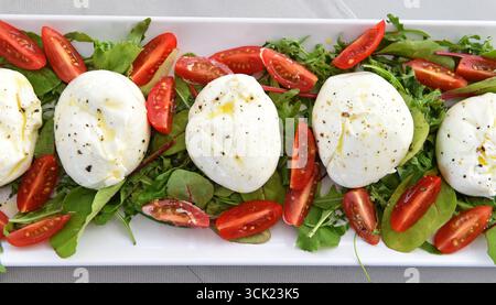 Ein lebendiger Blick von oben auf einen frischen Caprese-Salat, serviert auf einer weißen rechteckigen Platte. Das Gericht besteht aus mehreren weichen, cremigen Hügeln Burrata-Käses Stockfoto