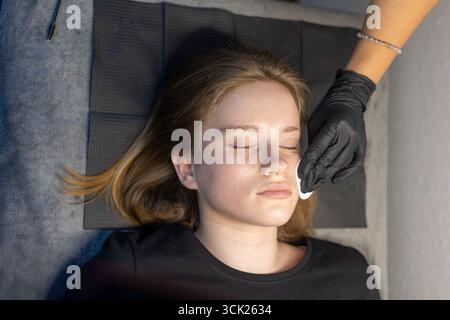 Eine junge Frau liegt mit geschlossenen Augen und erhält eine Gesichtsbehandlung. Eine Kosmetikerin, die schwarze Handschuhe trägt, reinigt ihr Gesicht mit einem Wattepad in einem Schönheitsbereich Stockfoto