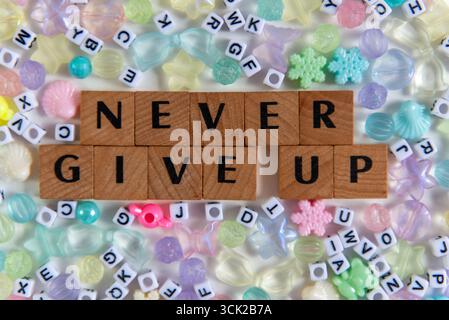 Inspirierendes Zitat „Never Gib up“, geschrieben mit Holzfliesen, umgeben von Pastellperlen und Plastikbuchstaben. Kreativer und motivierender, flacher Lehnstuhl Stockfoto