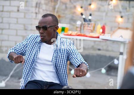 Schwarzer junger Erwachsener, der eine Sonnenbrille trägt und im Freien tanzt, festlicher Tisch mit Speisen und Getränken im Hintergrund Stockfoto