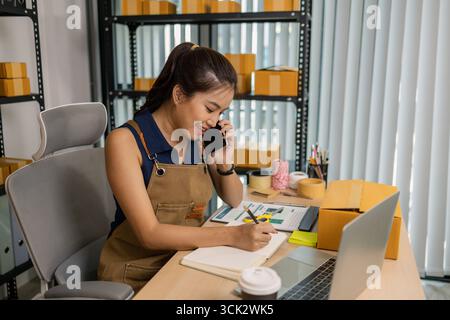 Lächelnde junge Unternehmerin, die vom Home Office aus auf dem Handy spricht und am Laptop arbeitet, die Kundenbestellungen für ihre Online-Kleidung verwaltet Stockfoto