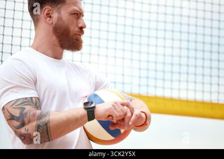 Ein bärtiger Mann mit Tätowierungen hält einen Volleyball, trägt ein weißes T-Shirt und eine Uhr. Draußen vor einem gelben Volleyballnetz zu stehen, ist die Szene verrückt Stockfoto